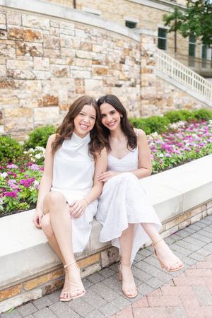 Two smiling young women in white dresses sit together on a stone ledge with colorful flowers blooming behind them.