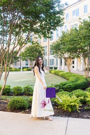 Young woman in white maxi dress holding purple graduation cap, smiling outdoors near university building.
