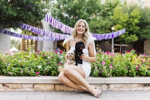 Smiling blonde woman in white dress sitting outdoors holding two small dogs with colorful flowers and festive bunting behind her.