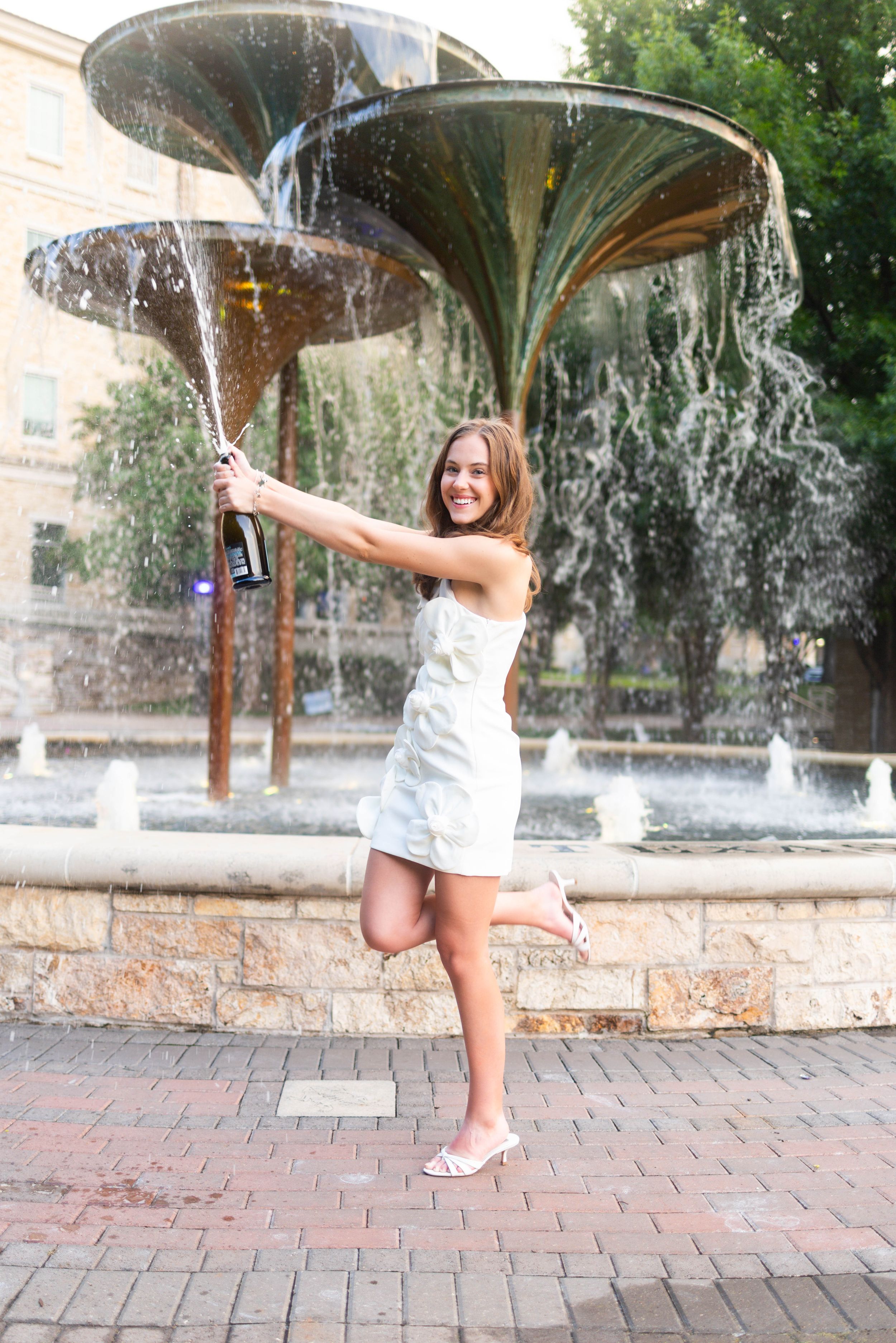 Young woman in white dress celebrating with champagne bottle at decorative fountain outdoors.