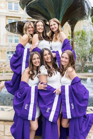 Six young women in purple TCU graduation gowns pose together smiling in front of a decorative fountain.