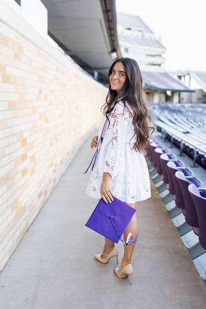 Smiling young woman in white dress holding purple graduation cap and tassel in stadium seating area.