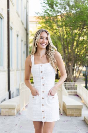 Smiling young woman in white button-front mini dress posing outdoors on a sunny day.