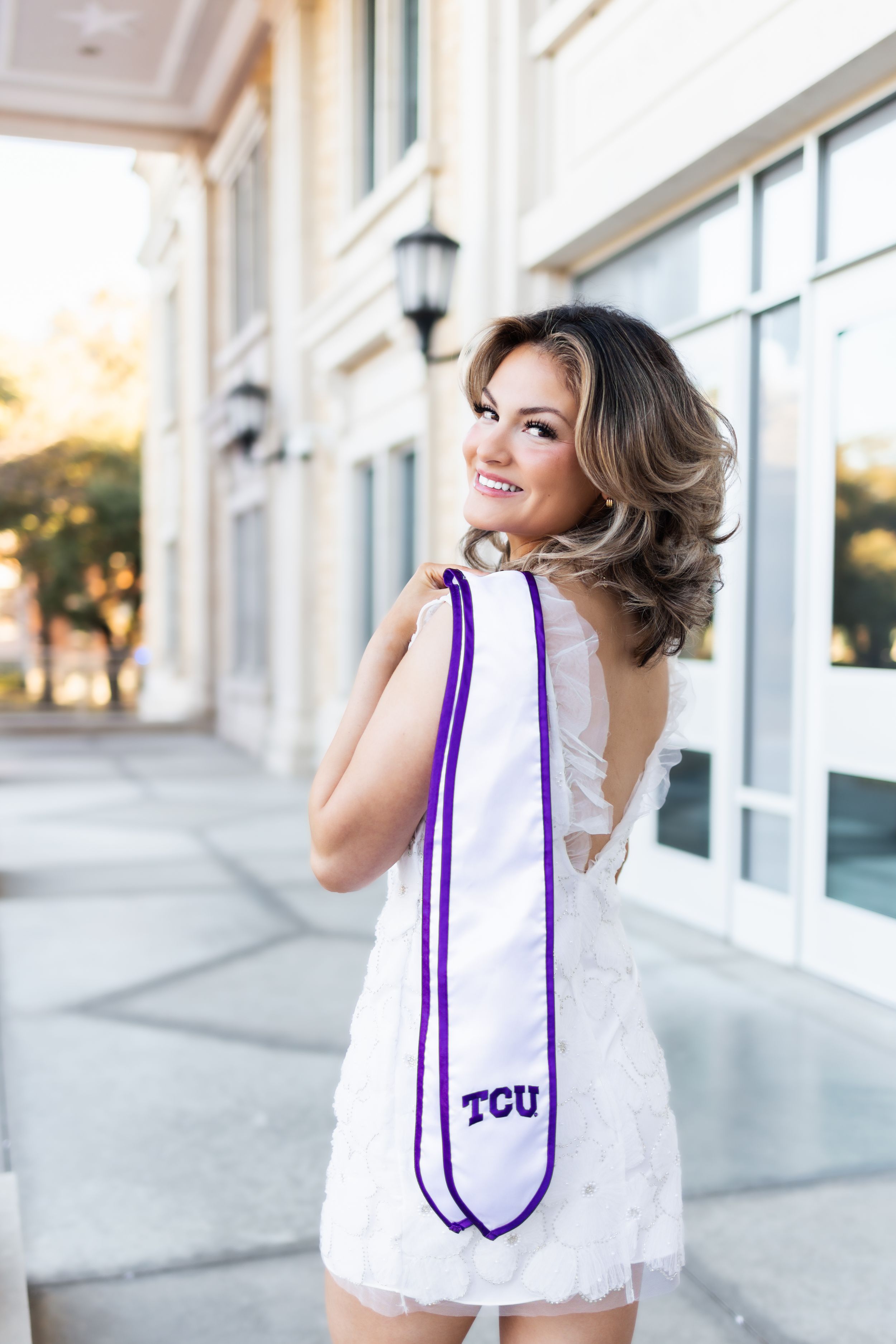 Smiling young woman in white dress holding a TCU graduation stole over her shoulder outdoors.