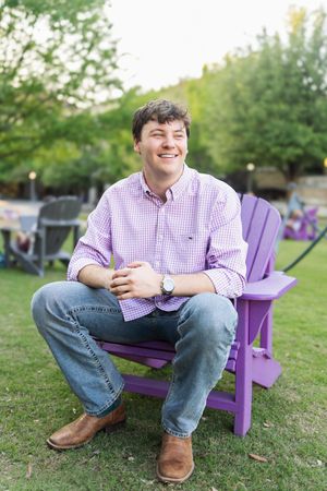 Young man smiling in purple Adirondack chair outdoors, wearing pink gingham shirt, jeans, and brown boots.