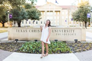 Young woman in white dress posing in front of Texas Christian University founded 1873 sign on campus.