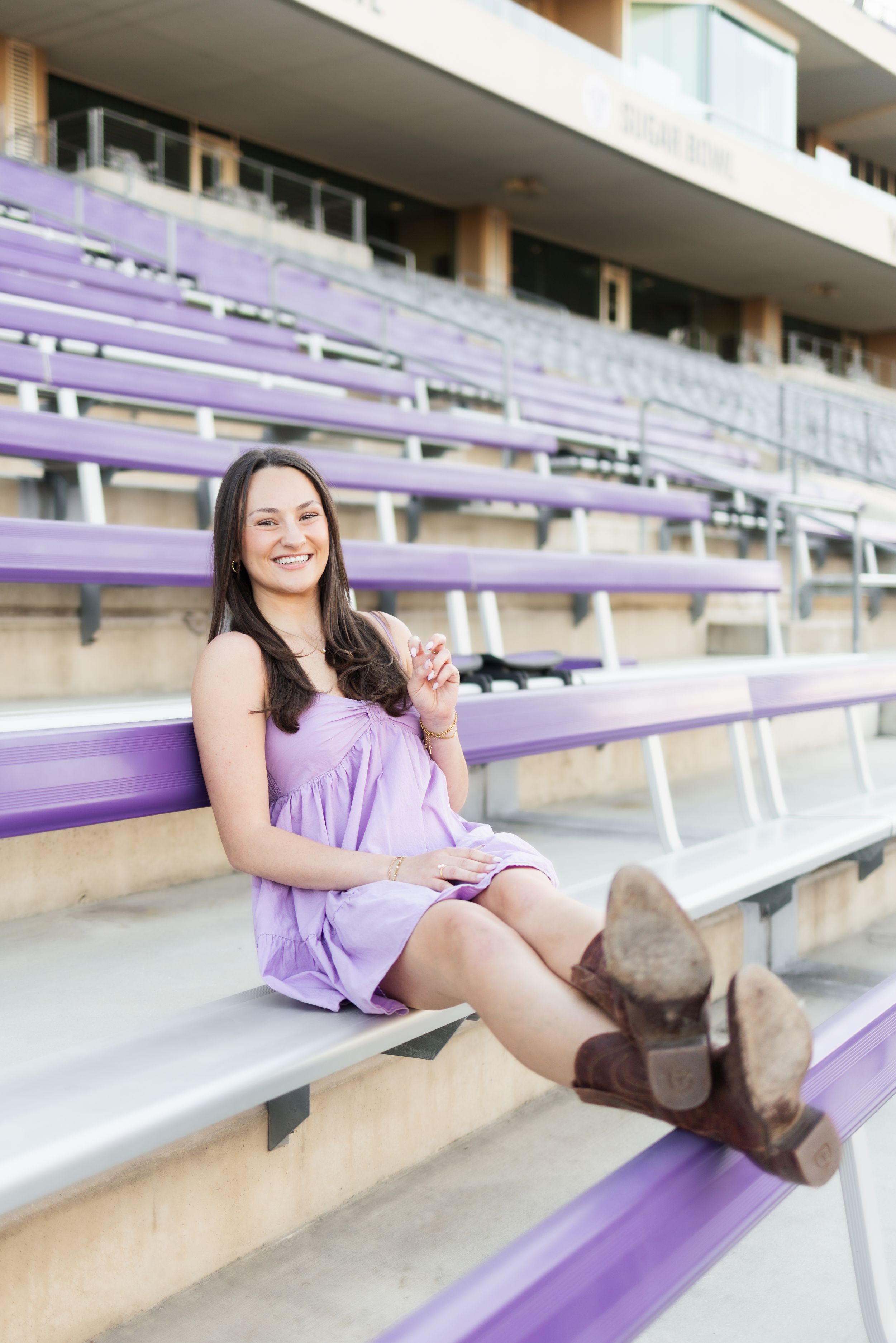 Young woman in purple dress and cowboy boots sitting on bleachers, smiling at camera.