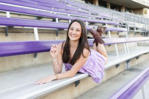 Young woman in purple outfit smiling while posing on purple stadium bleachers outdoors.