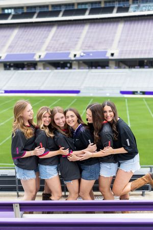Six young women in matching black jerseys and denim shorts pose together smiling in a purple stadium.