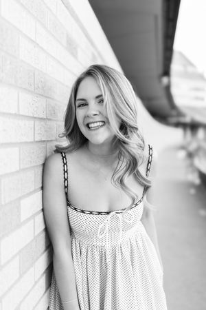 Smiling young woman with long blonde hair leaning against a brick wall in a polka dot sundress, black and white photo.