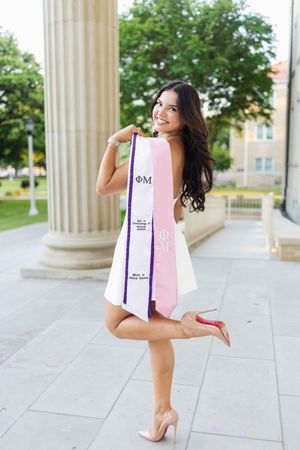 Smiling young woman in white dress holding Phi Mu graduation sash, posing outdoors near columns on a college campus.