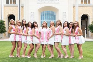 Group of young women in matching white dresses and pink sashes posing together outdoors in front of a building.