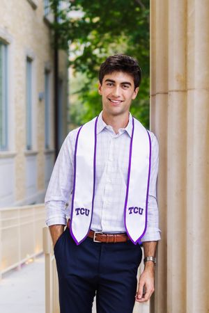 Young man smiling in TCU graduation stole and dress shirt, posing between stone columns outdoors.