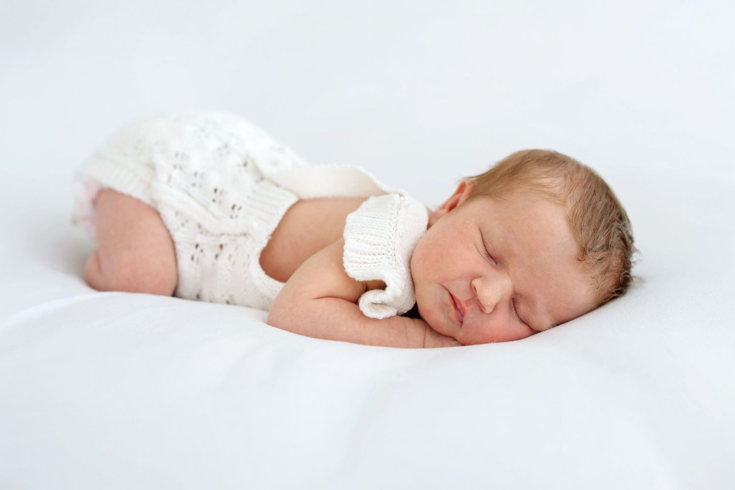 Peaceful newborn baby sleeping on white blanket wearing a cozy white knit outfit and hat.