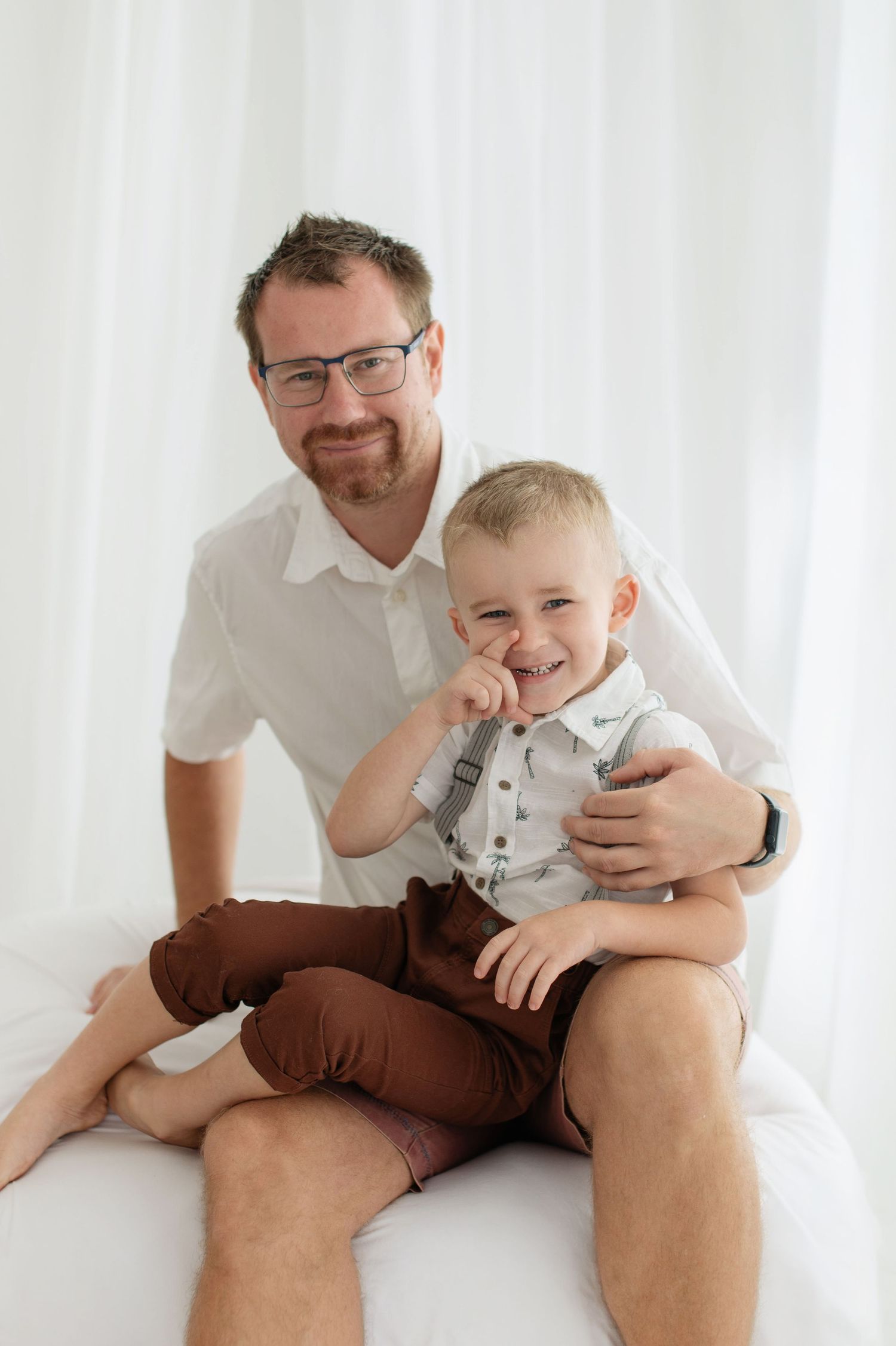 Father with glasses smiles holding laughing toddler boy in suspenders during white studio family photo session.