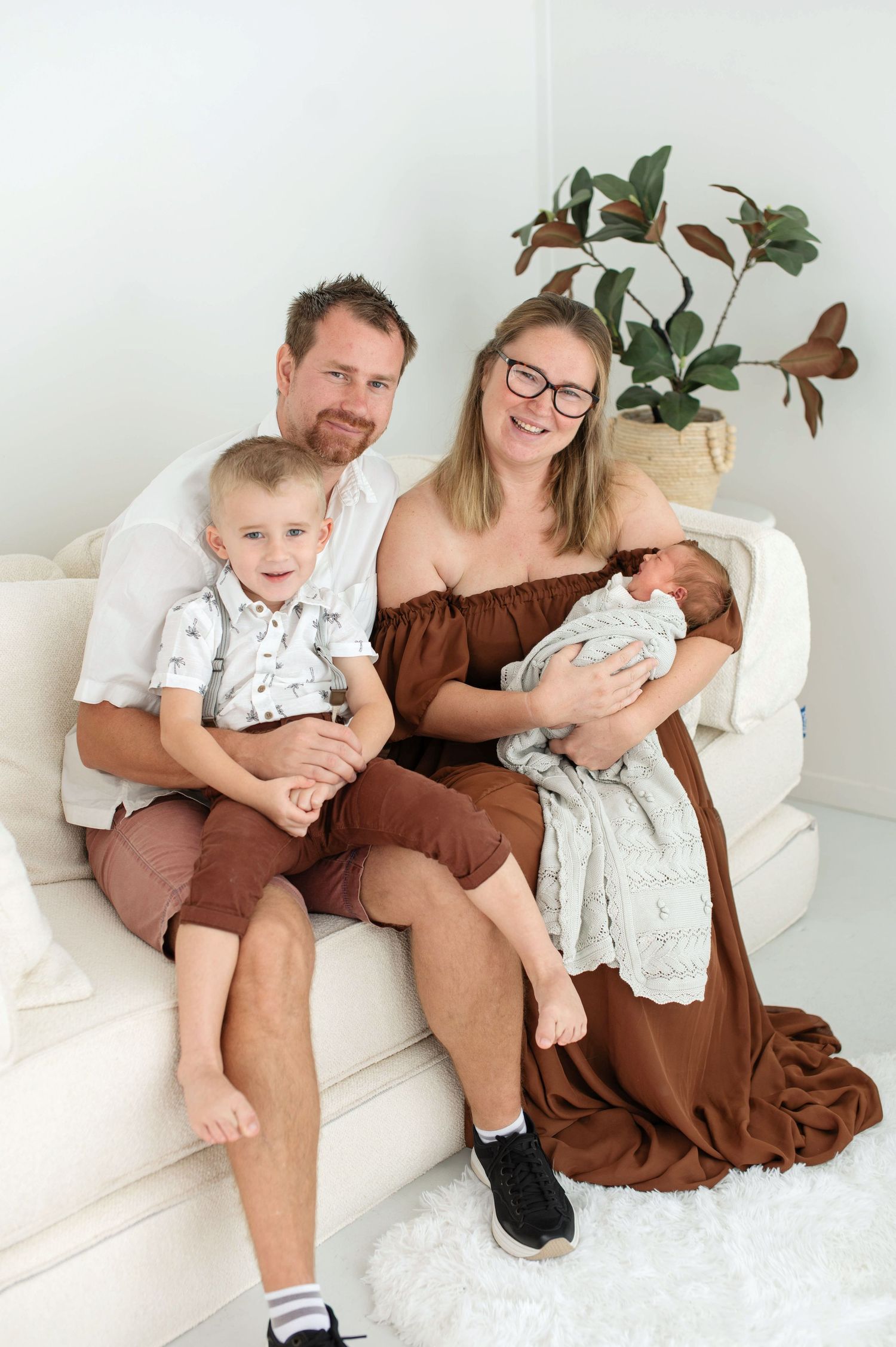 Happy family of four sitting on white couch, parents with toddler son and newborn baby, wearing coordinated brown and white outfits.