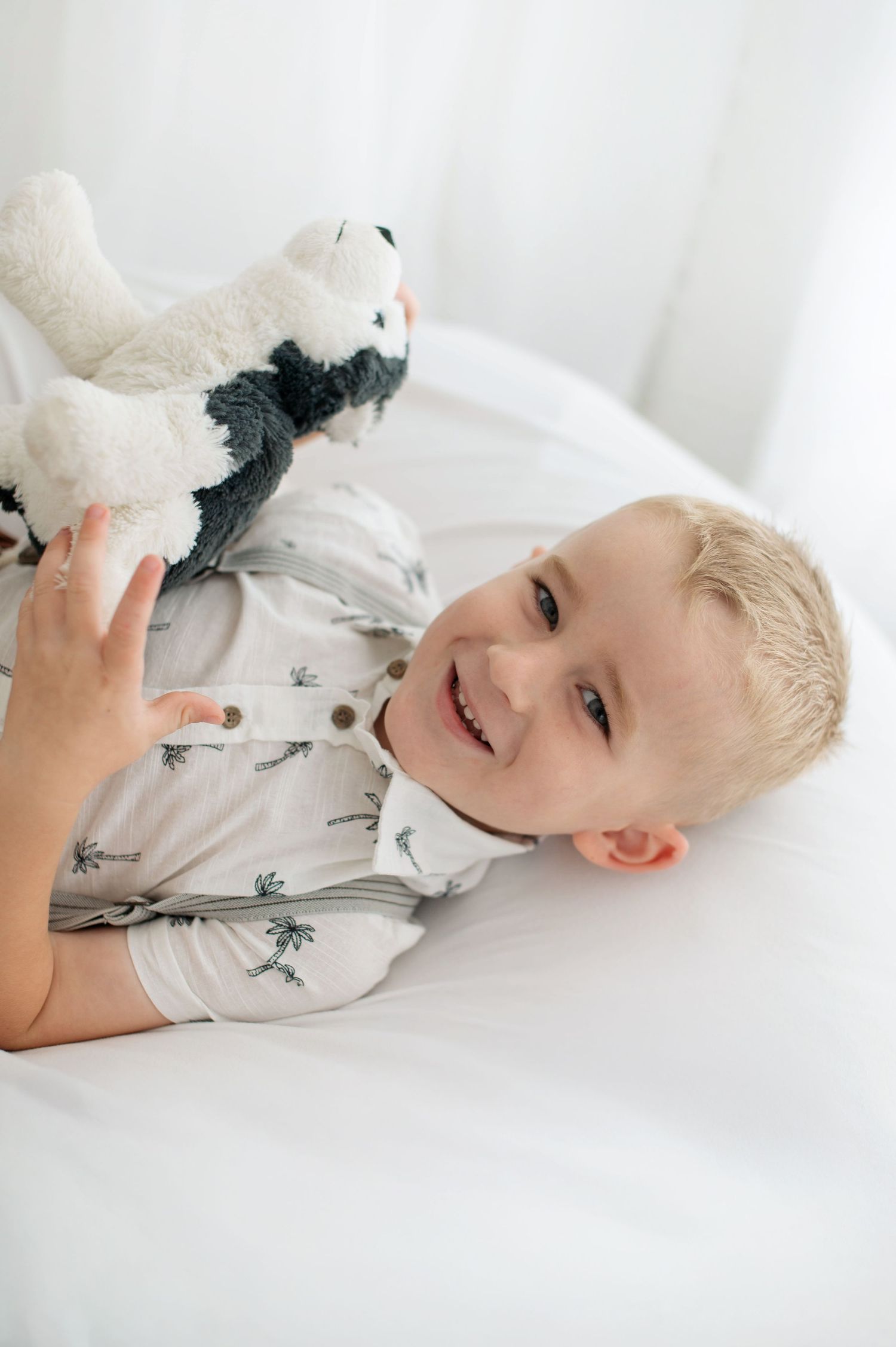 Smiling blonde toddler boy lying on white bed holding a stuffed animal toy.