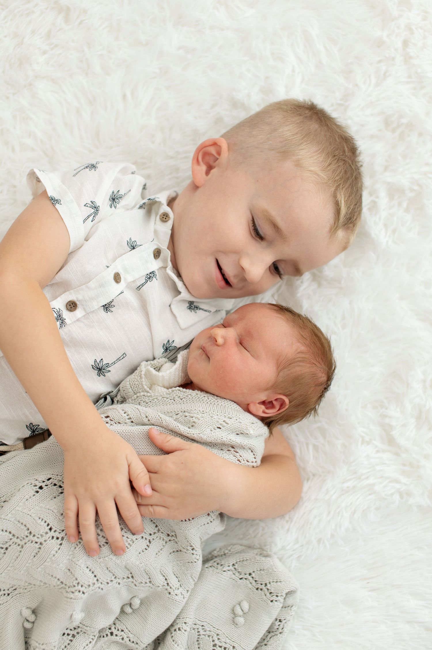 Young blonde boy smiling at sleeping newborn baby sibling on white fluffy blanket.