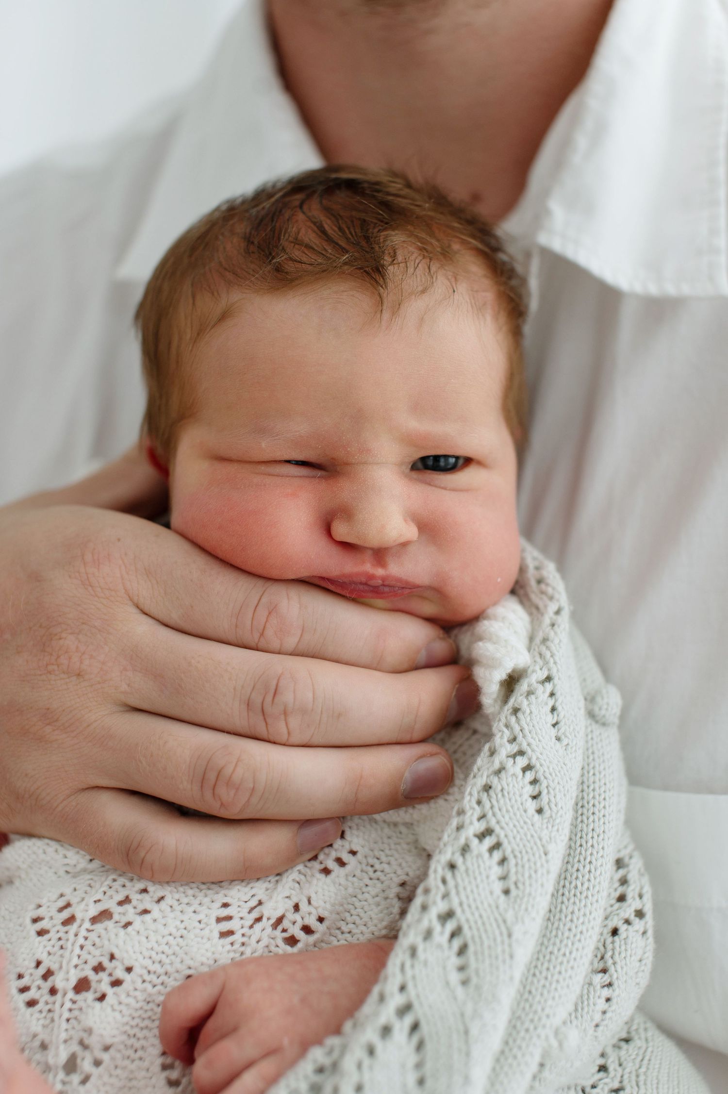 Newborn baby wrapped in white knit blanket, cradled in adult hands against white shirt.