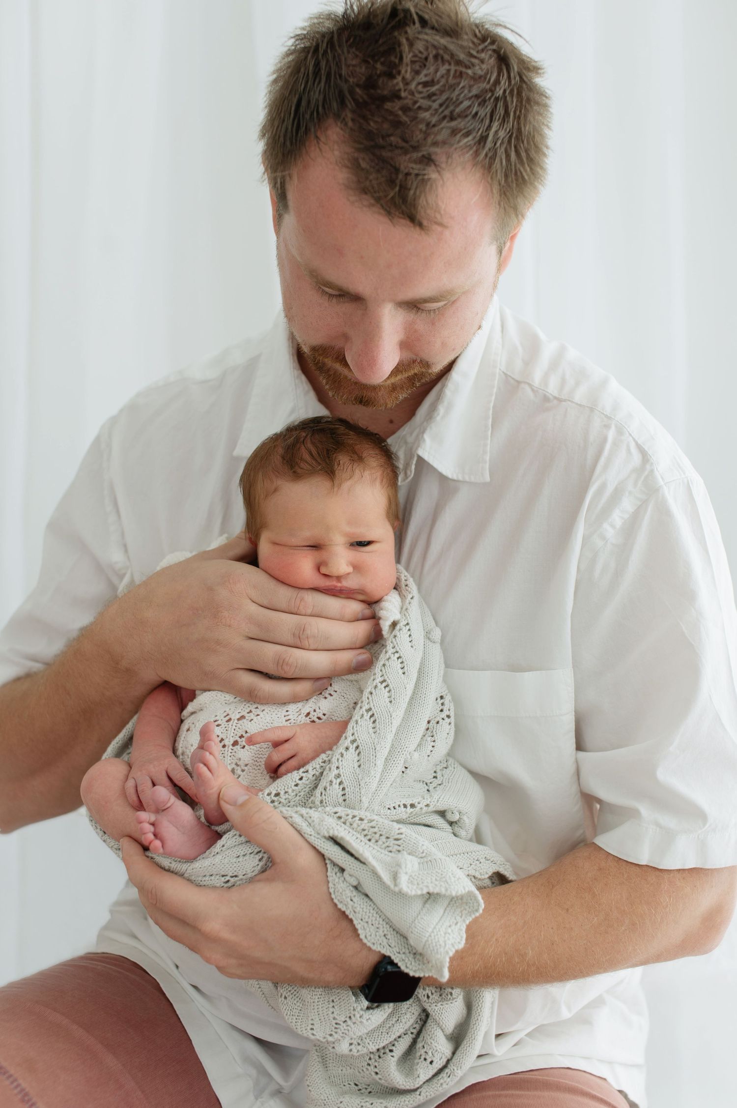 Father in white shirt tenderly holds sleeping newborn baby wrapped in knit blanket during studio session.