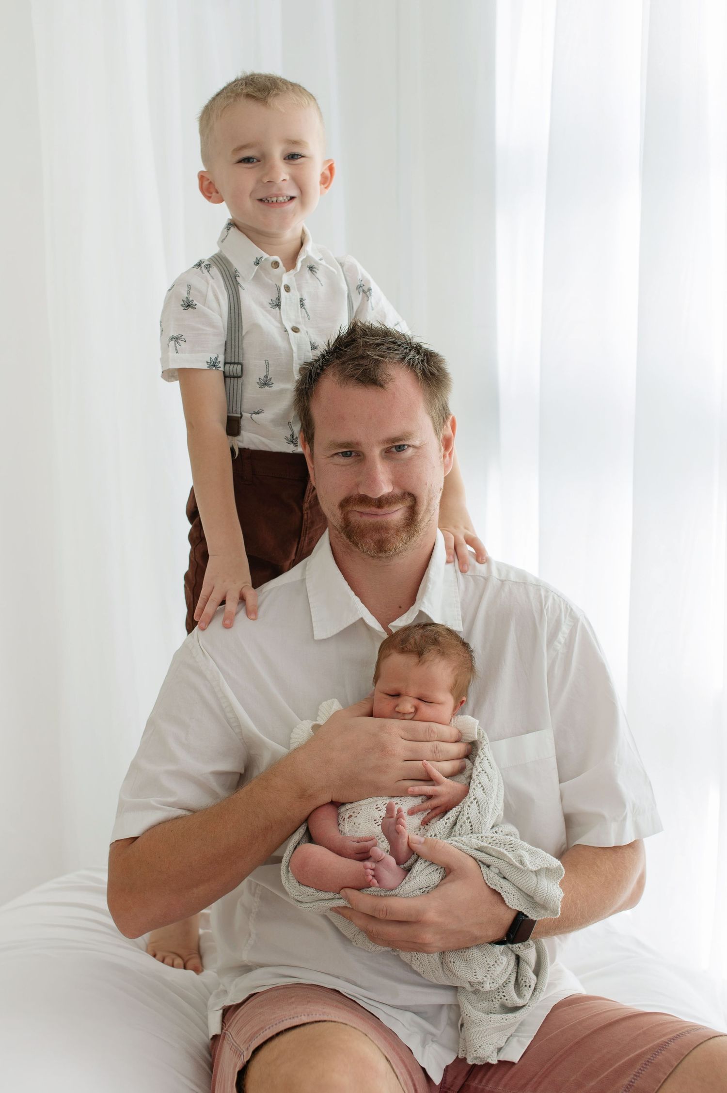 Father sitting with newborn baby while young blonde son stands behind him smiling.