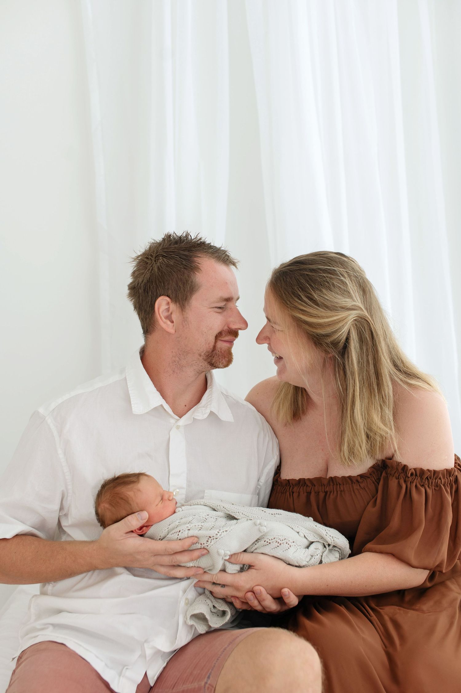 Loving couple smiling at each other while tenderly holding their swaddled newborn baby in a bright white studio.