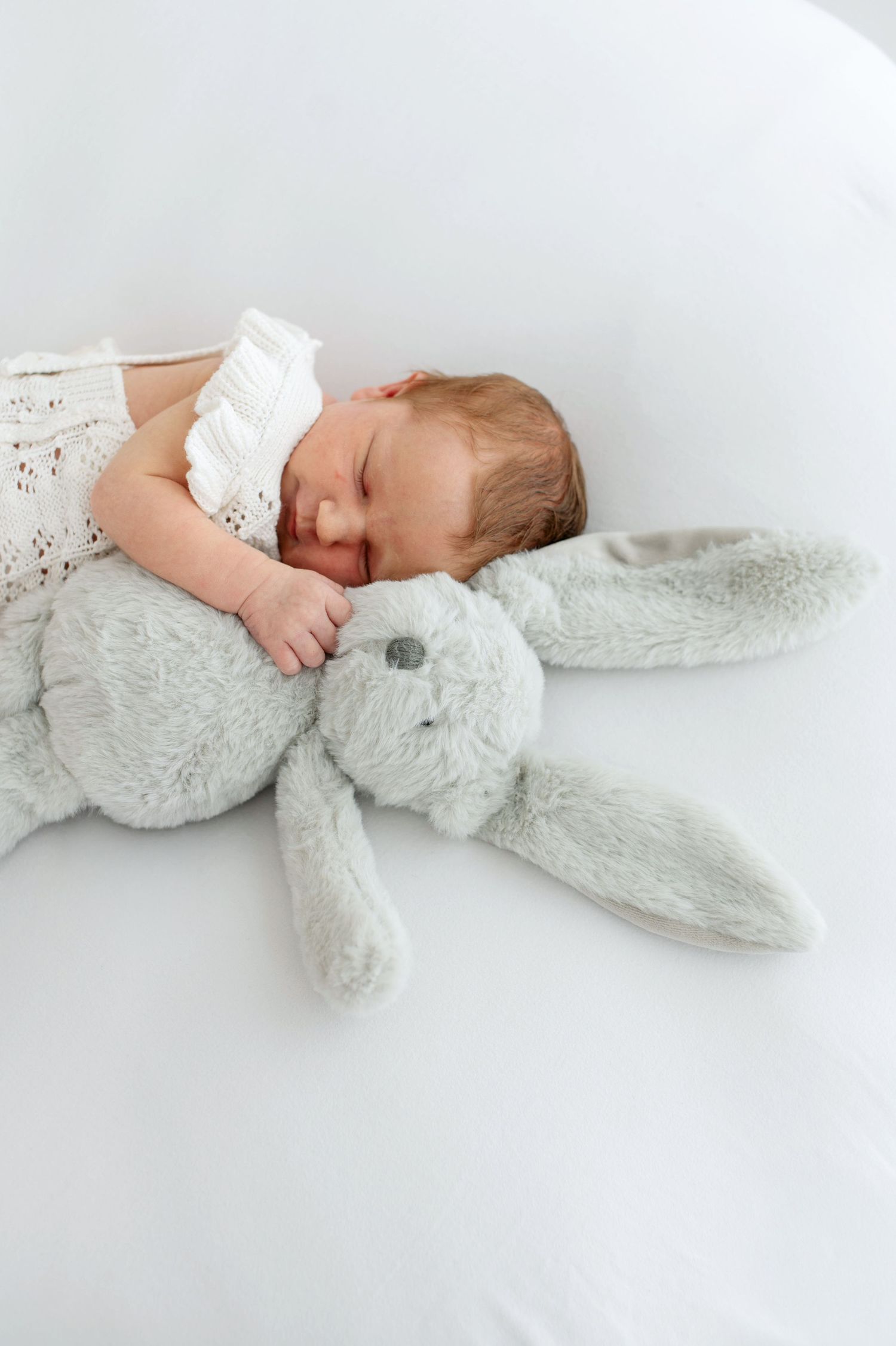 Newborn baby sleeping peacefully on white bedding while cuddling a soft grey plush bunny toy.