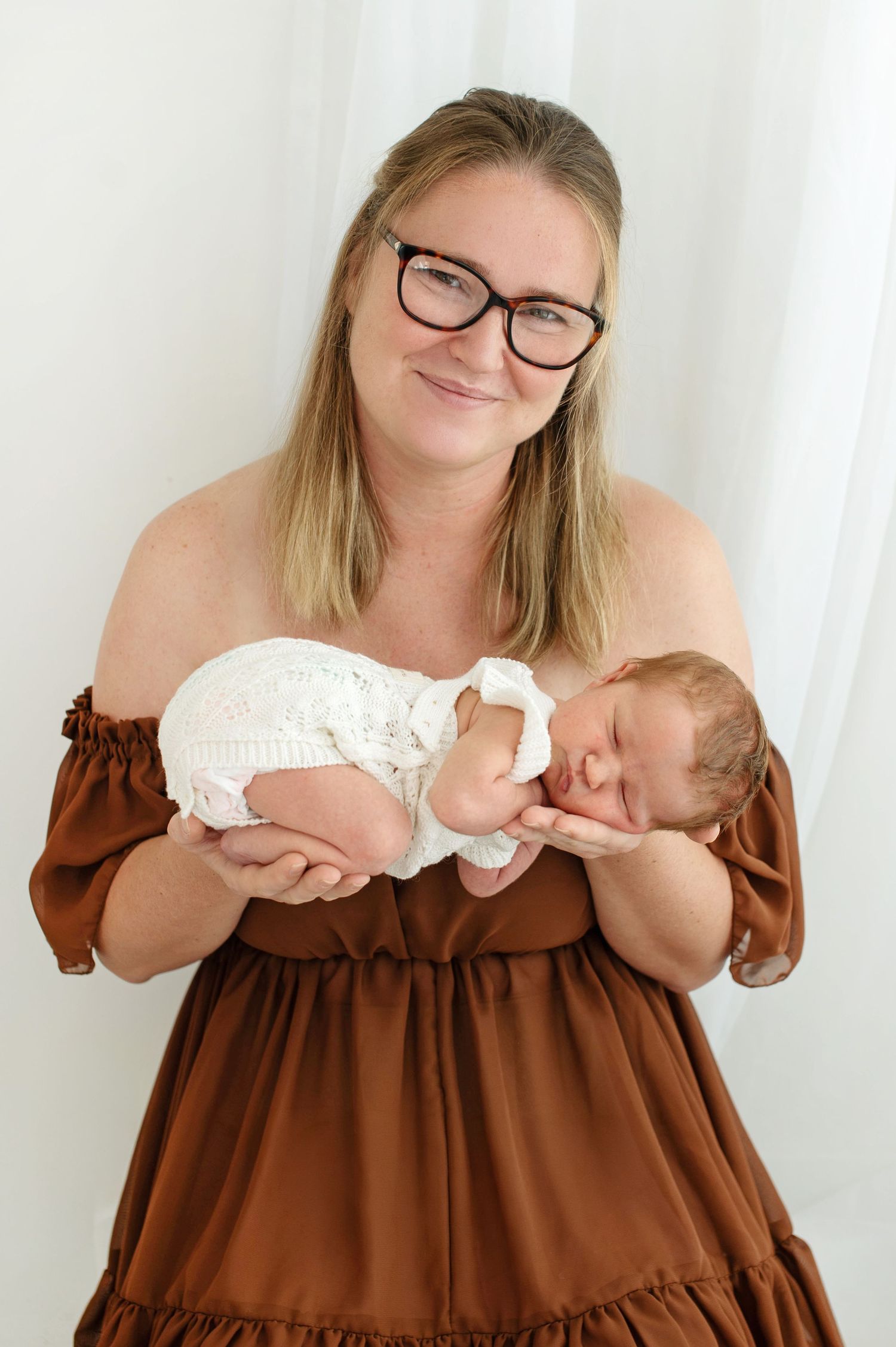 Smiling mother in brown off-shoulder dress holding sleeping newborn baby in white crochet outfit.