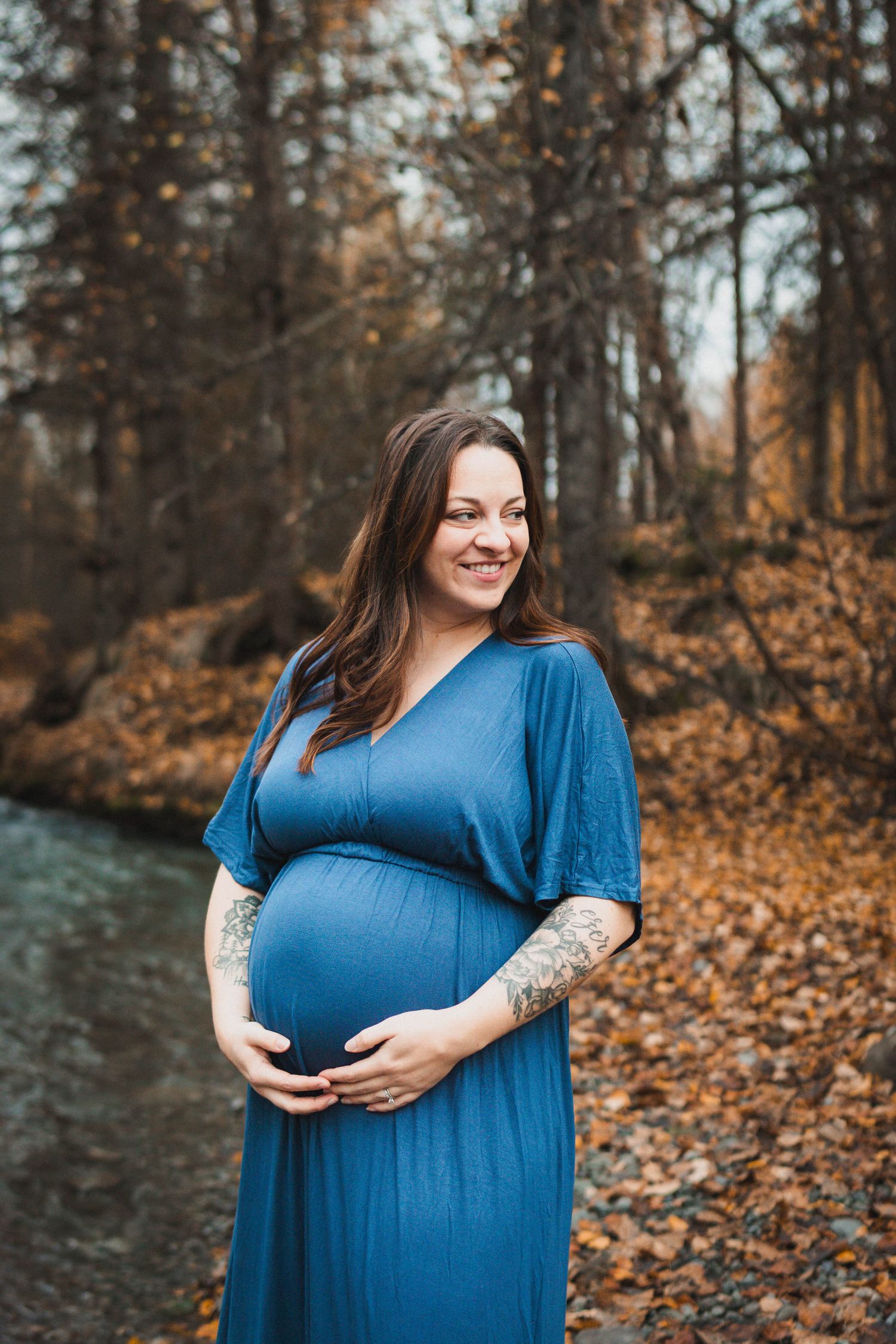 Smiling pregnant woman in blue dress holding baby bump outdoors in autumn forest setting.