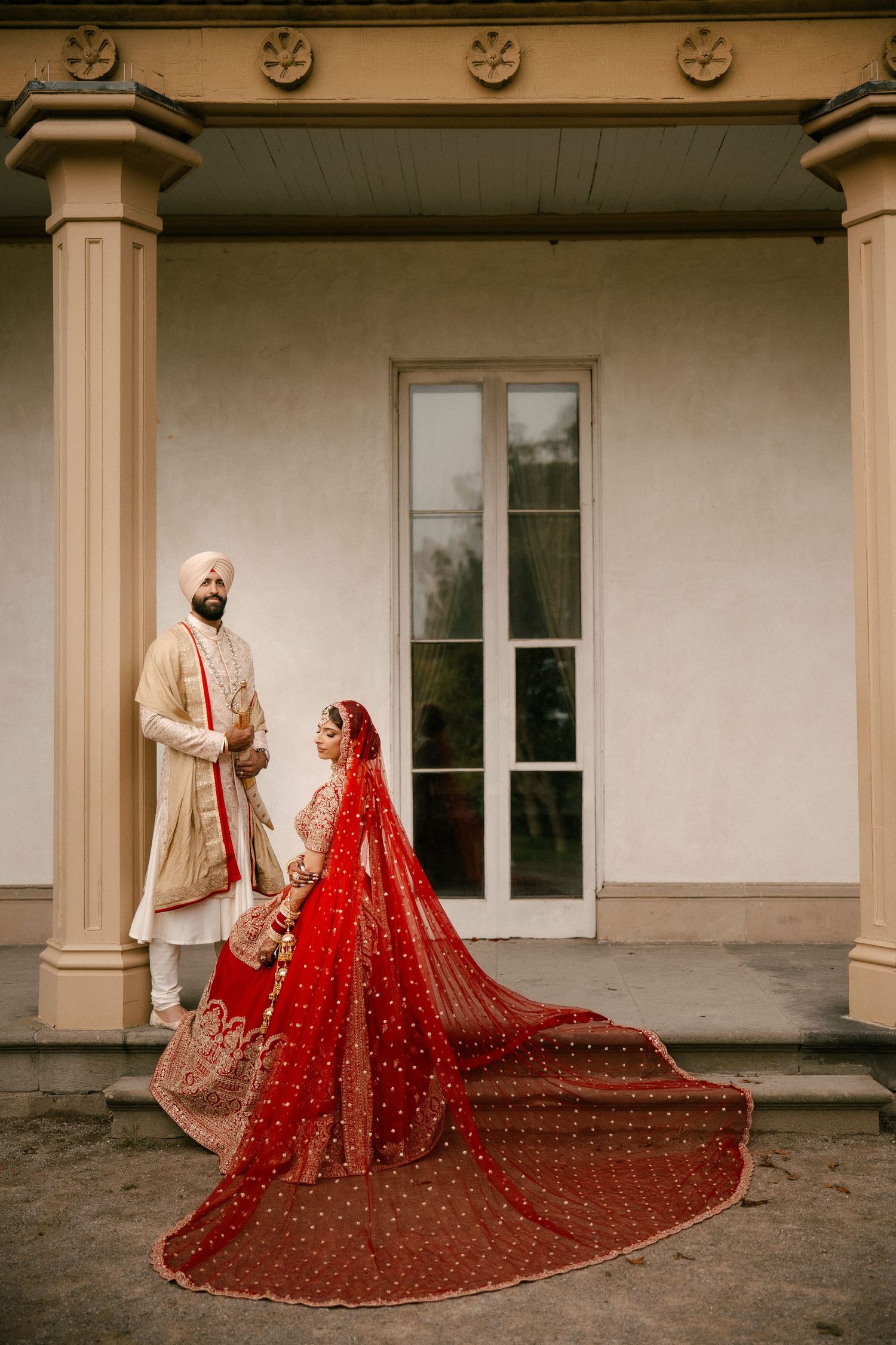 Indian bride in red lehenga and groom in white sherwani walking through golden meadow during outdoor wedding photography.