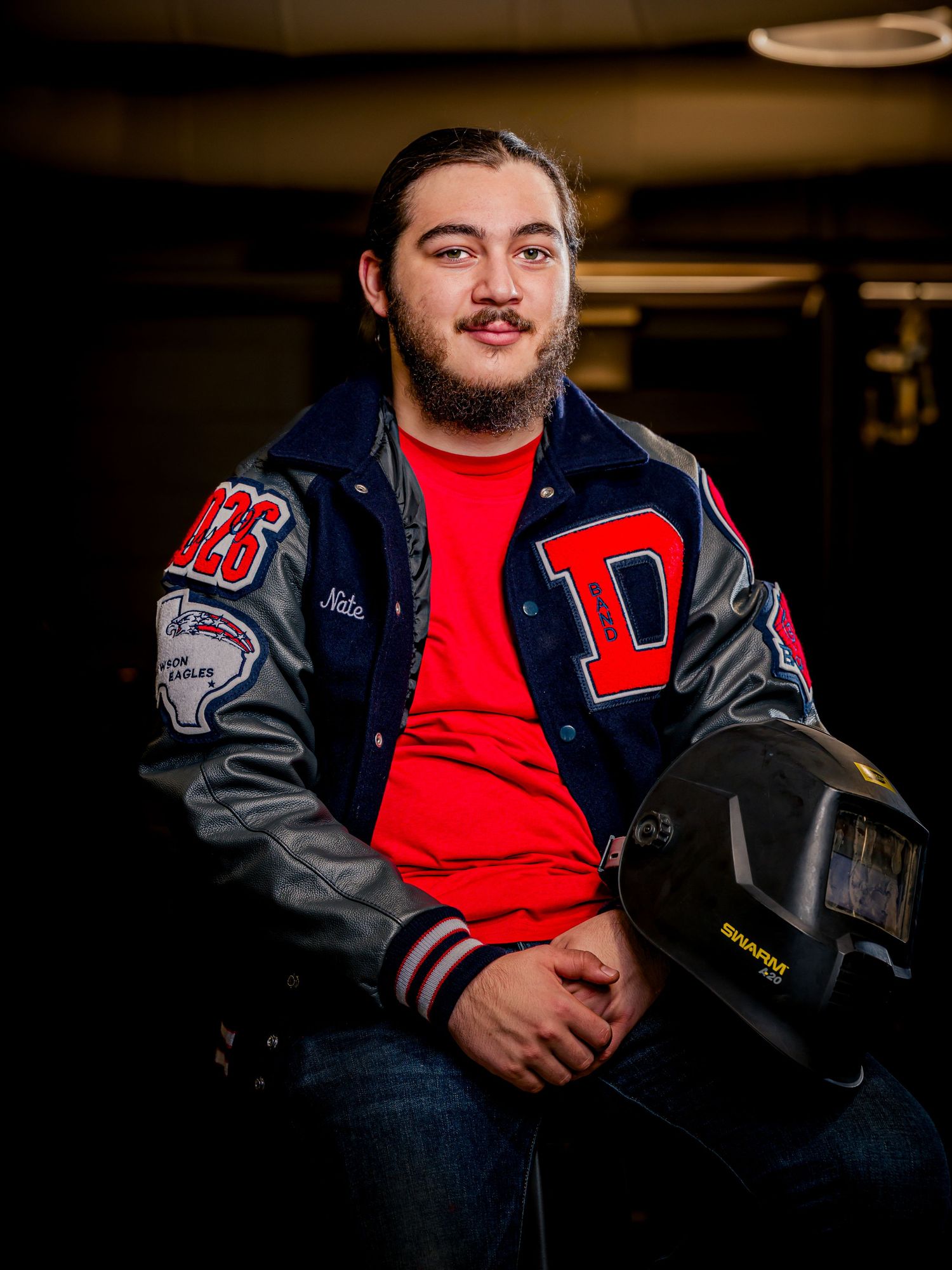 Young man in a letterman jacket poses with a welding helmet in a dark industrial workshop setting.