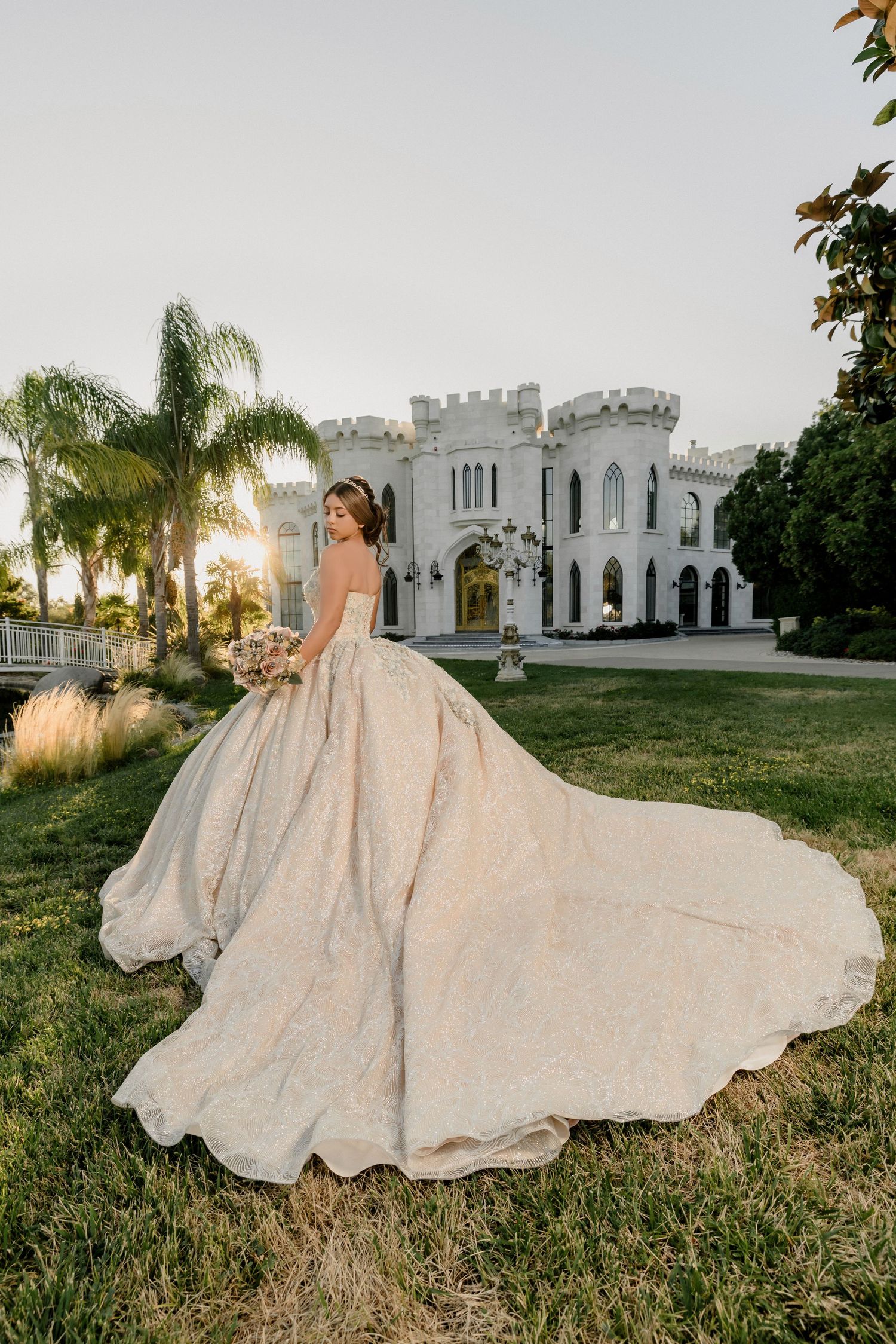Bride in a flowing ballgown with long train poses before a castle venue at sunset, holding a bouquet.