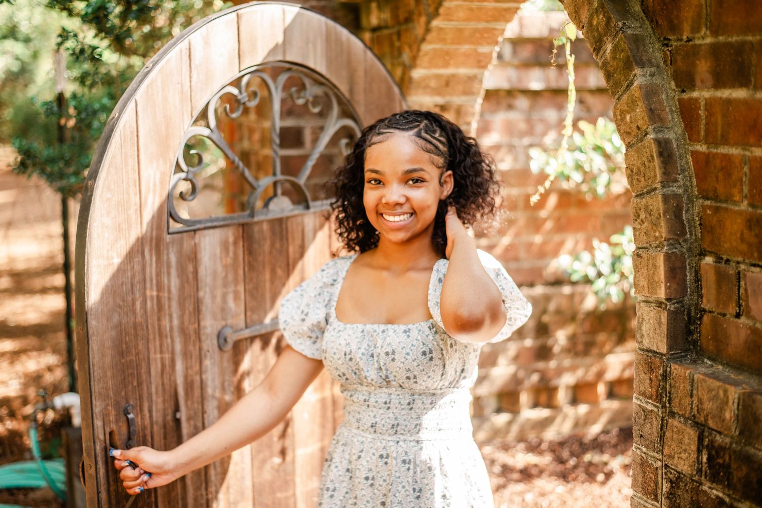 Young Black girl in floral white dress smiling by rustic wooden garden gate with brick arch.