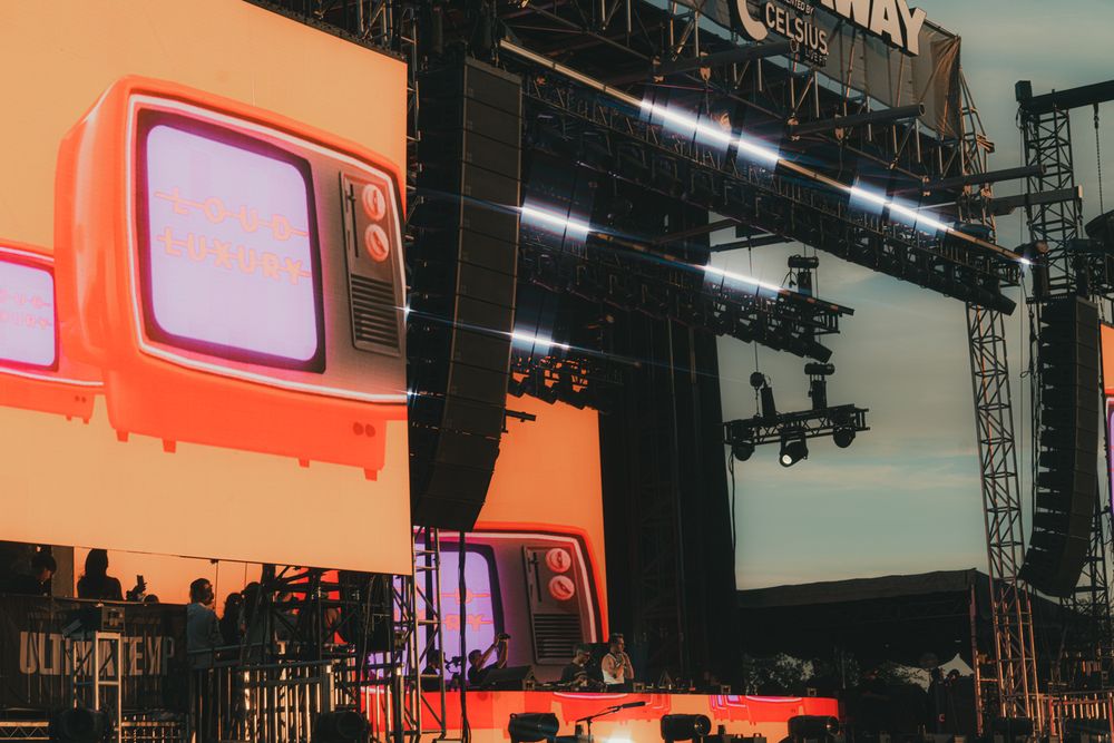 Large LED screens and stage lighting rigs at an outdoor music festival concert venue at sunset.