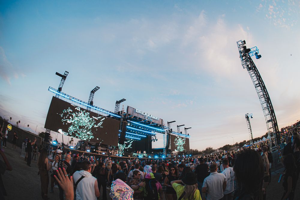 Large outdoor music festival crowd gathered at dusk before a massive illuminated stage with confetti and lights.