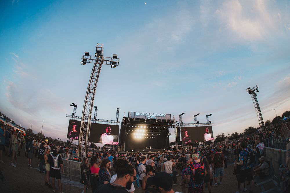 Crowd gathered at an outdoor music festival stage with large LED screens at dusk under a blue sky.