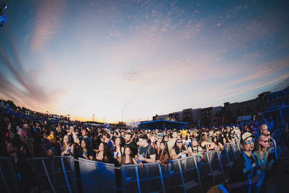 Large outdoor music festival crowd at sunset with colorful sky and stage lighting illuminating excited fans.