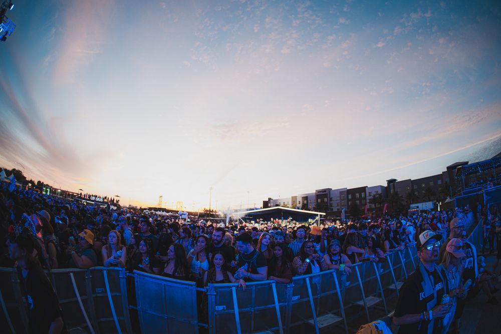 Large outdoor music festival crowd at sunset with colorful stage lights and clear sky backdrop.