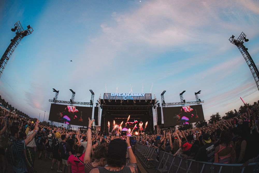 Massive outdoor music festival crowd cheering before a large illuminated stage at dusk with colorful lights.
