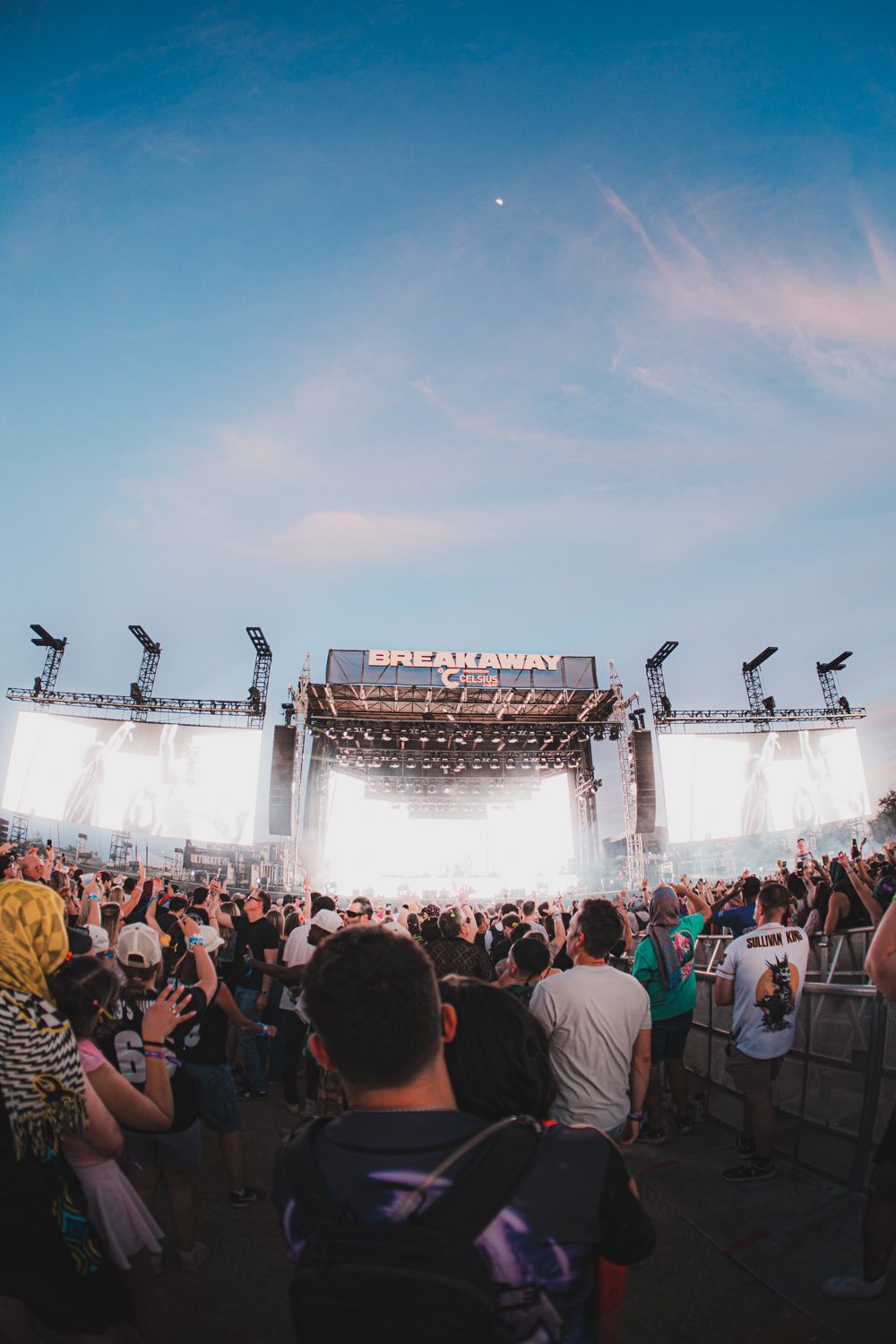 Large crowd at Breakaway Music Festival facing a brightly lit stage at dusk under a clear blue sky.