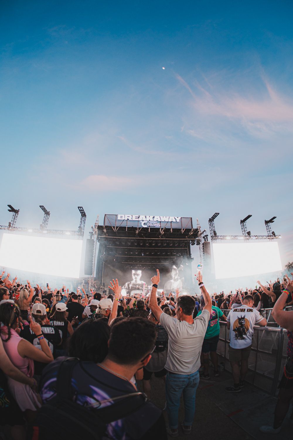 Crowd of fans with raised hands at Breakaway Music Festival outdoor stage with bright lights at dusk.