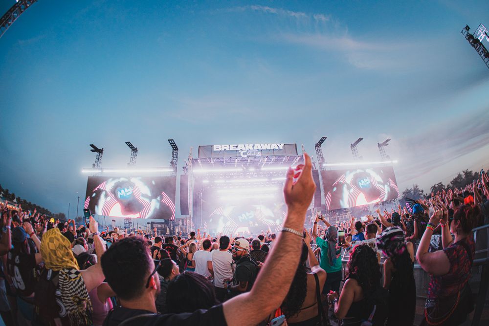 Crowd enjoying Breakaway Music Festival with hands raised at an outdoor stage lit with colorful lights at dusk.