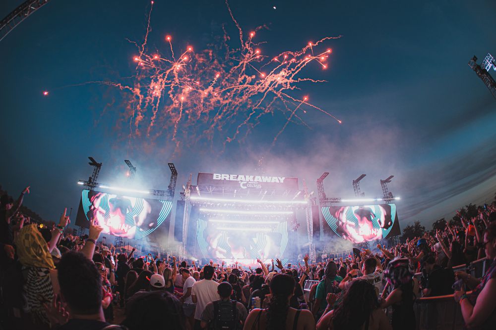 Breakaway music festival stage with fireworks exploding above a massive crowd at dusk.