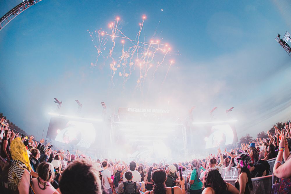 Crowd at outdoor music festival with bright stage lights and colorful fireworks exploding in the sky above.