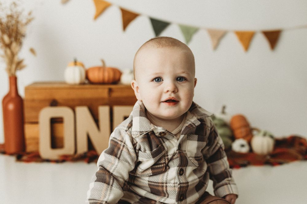Baby in plaid shirt sits before ONE sign with fall pumpkins and bunting for a first birthday cake smash photo.