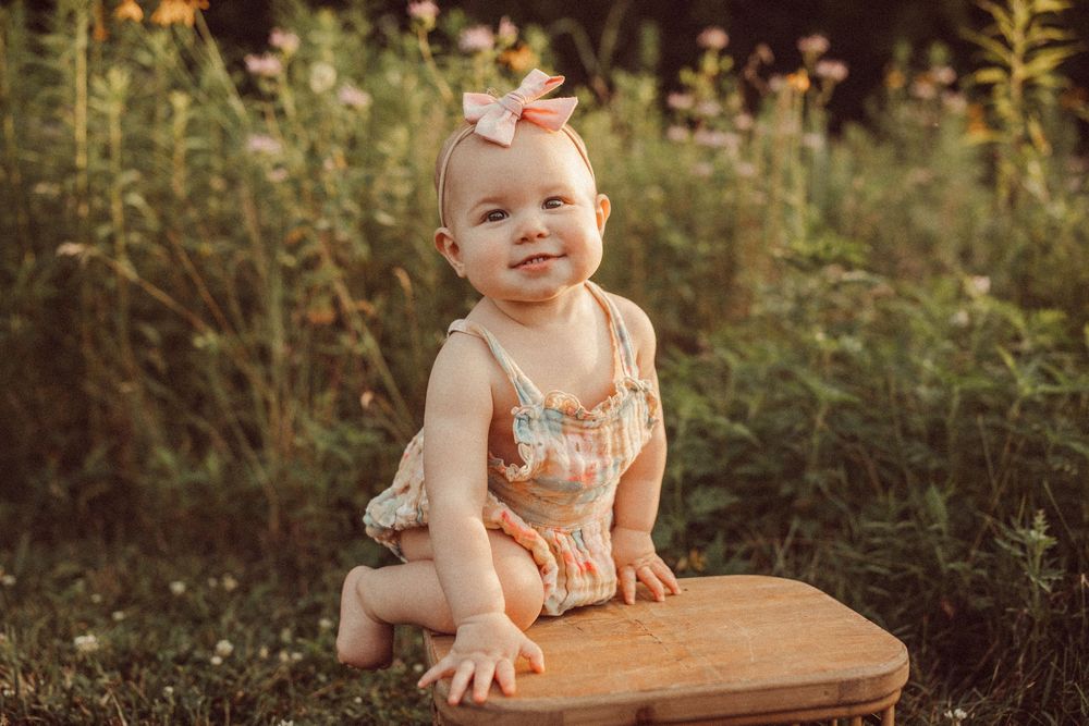 Smiling baby girl in floral romper and bow headband sitting outdoors in a sunny garden setting.