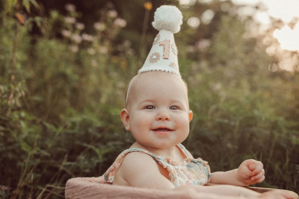 Smiling baby wearing a party hat sitting outdoors in a wooden box surrounded by greenery during a first birthday photoshoot.