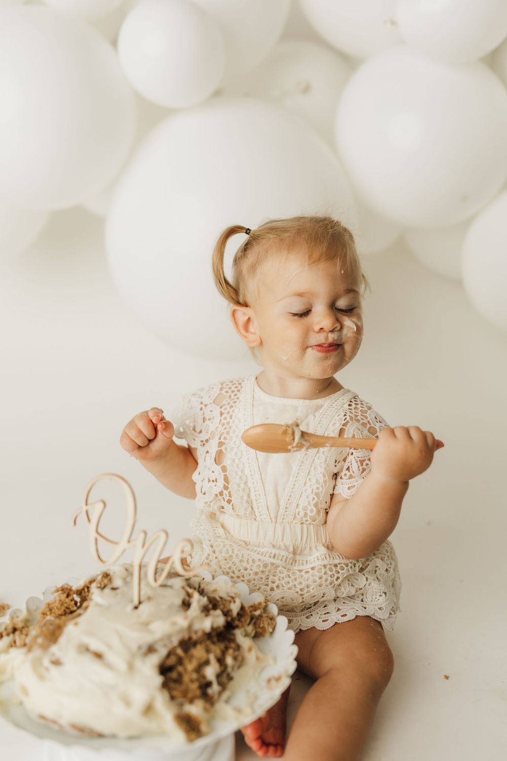Toddler girl in lace romper smashing her first birthday cake with a wooden spoon, white balloons in background.