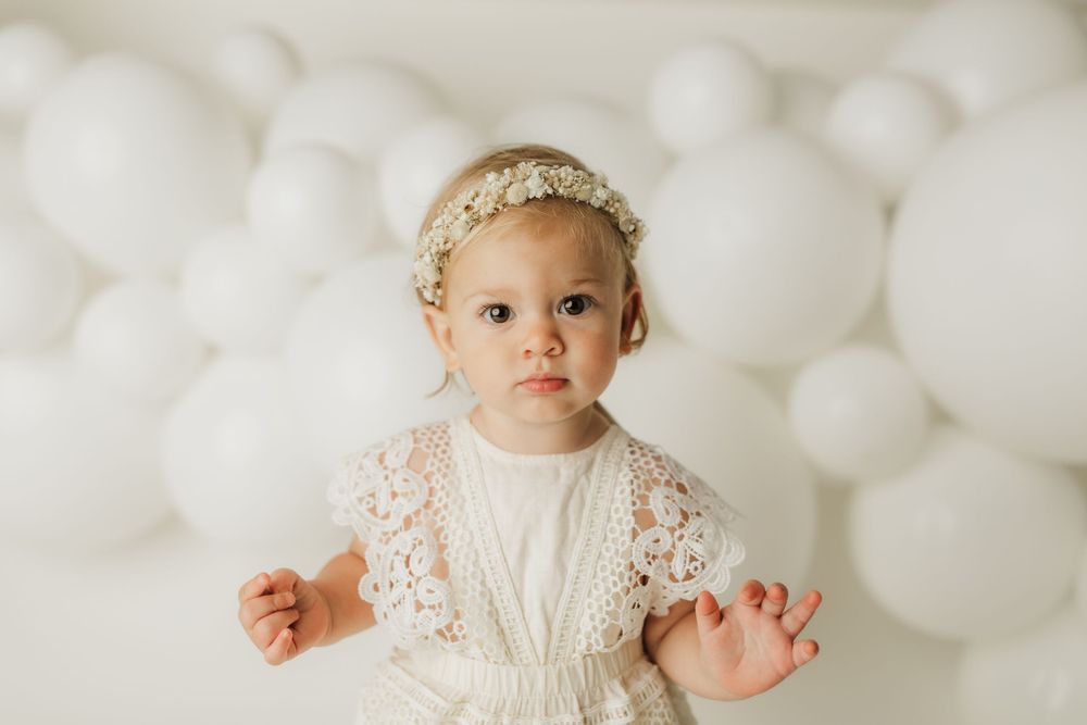 Adorable baby girl in white lace dress and floral headband poses in front of white balloon backdrop.