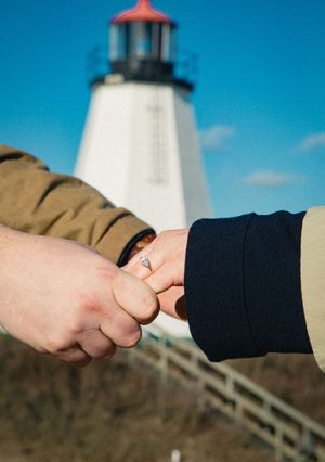Close-up of holding hands with engagement ring in front of lighthouse.